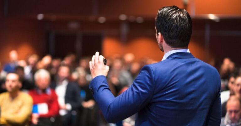 A speaker in a blue suit addresses an attentive audience during a presentation, illustrating the fundamentals of public speaking, including engaging delivery and audience connection.