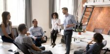 A group of professionals engaged in a lively discussion in a modern office setting with a mix of natural light and exposed brick walls. A man is leading the conversation, suggesting a collaborative coaching environment. The image illustrates a supportive and interactive atmosphere, ideal for those learning how to start a coaching business.