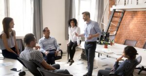 A group of professionals engaged in a lively discussion in a modern office setting with a mix of natural light and exposed brick walls. A man is leading the conversation, suggesting a collaborative coaching environment. The image illustrates a supportive and interactive atmosphere, ideal for those learning how to start a coaching business.