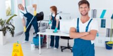 A team of professional cleaners in blue uniforms working in a modern office space, with a confident man in the foreground and others cleaning in the background. This image represents the teamwork and dedication needed when starting a home services business.