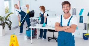 A team of professional cleaners in blue uniforms working in a modern office space, with a confident man in the foreground and others cleaning in the background. This image represents the teamwork and dedication needed when starting a home services business.