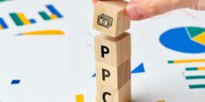 Wooden blocks spelling PPC with a money symbol on top, placed on charts and graphs, symbolizing pay-per-click (PPC) advertising and financial growth. A hand places the money block, representing PPC strategy management.