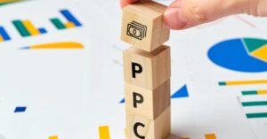 Wooden blocks spelling PPC with a money symbol on top, placed on charts and graphs, symbolizing pay-per-click (PPC) advertising and financial growth. A hand places the money block, representing PPC strategy management.