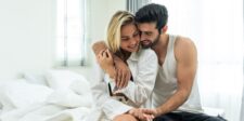 A couple sits closely on the bed, smiling and embracing each other in a light-filled bedroom, representing intimacy and connection. Their comfortable closeness reflects the emotional and intellectual bond between them.