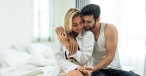 A couple sits closely on the bed, smiling and embracing each other in a light-filled bedroom, representing intimacy and connection. Their comfortable closeness reflects the emotional and intellectual bond between them.