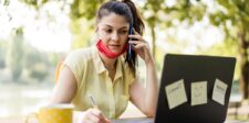 Solopreneur working outdoors, multitasking with a phone call and a laptop, while taking notes. A face mask hangs loosely around the neck, and a coffee cup is placed on the table beside the laptop.