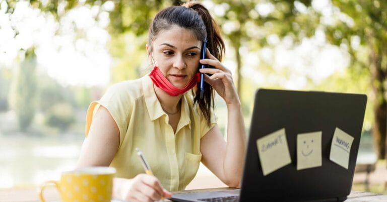 Solopreneur working outdoors, multitasking with a phone call and a laptop, while taking notes. A face mask hangs loosely around the neck, and a coffee cup is placed on the table beside the laptop.