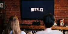 Two people sitting on a couch watching Netflix on a TV in a cozy living room with brick walls. The scene suggests a relaxed environment, perfect for viewing entrepreneur-themed movies available on Netflix.