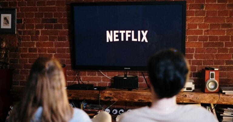 Two people sitting on a couch watching Netflix on a TV in a cozy living room with brick walls. The scene suggests a relaxed environment, perfect for viewing entrepreneur-themed movies available on Netflix.
