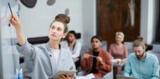 Professional woman leading a coaching session, writing on a whiteboard, with a group of attentive individuals in the background. This image highlights a collaborative learning environment, ideal for coaching and counseling settings.