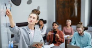 Professional woman leading a coaching session, writing on a whiteboard, with a group of attentive individuals in the background. This image highlights a collaborative learning environment, ideal for coaching and counseling settings.