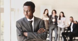 Man in a suit standing alone near a window, appearing deep in thought while a group of people socialize in the background, symbolizing the lonely path.