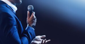 Close-up of a speaker holding a microphone, gesturing with one hand, illustrating the basics of good speaking, including connection, clarity, and confidence in public speaking.