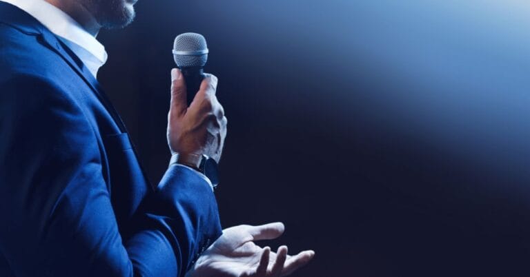 Close-up of a speaker holding a microphone, gesturing with one hand, illustrating the basics of good speaking, including connection, clarity, and confidence in public speaking.