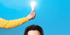 A hand holding a glowing light bulb above a person's head, symbolizing the moment of inspiration and creativity, with a bright blue background representing fresh ideas. This image reflects the journey from idea to invention.