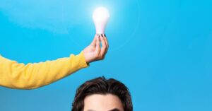 A hand holding a glowing light bulb above a person's head, symbolizing the moment of inspiration and creativity, with a bright blue background representing fresh ideas. This image reflects the journey from idea to invention.