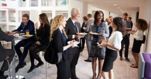 Professionals networking and socializing at a business event, holding plates with refreshments and engaging in conversations, illustrating a typical business networking event setting.