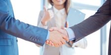 Two business professionals in suits shaking hands, with a woman in the background giving a thumbs-up, symbolizing agreement and successful collaboration.