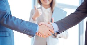 Two business professionals in suits shaking hands, with a woman in the background giving a thumbs-up, symbolizing agreement and successful collaboration.