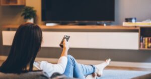 A person sitting on a couch in a cozy living room, holding a remote control and facing a large flat-screen TV on a modern entertainment unit. The relaxed setting suggests enjoying TV shows or movies at home.