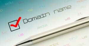 A metallic pen resting below the text 'Domain name' with a red checkmark in a checkbox, surrounded by colorful domain extensions like .com, .edu, and .us, symbolizing domain registration.