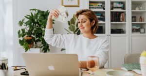 A woman sitting at her desk with a laptop, casually holding a stack of cash, showcasing financial confidence and the traits of a money magnet. The background includes bookshelves and plants, adding a sense of balance and success.