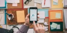 15 LinkedIn Alternatives for Professional Networking Overhead view of a desk filled with colorful folders, sticky notes, glasses, and office supplies, while a person holds a smartphone displaying data analytics graphs and charts. This image represents the organized, data-driven approach that professionals can achieve using a LinkedIn alternative for networking and productivity.