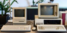 Two vintage desktop computers with monochrome monitors and bulky keyboards displayed on a desk, representing the technological era when the oldest websites were created, with a retro ambiance complemented by a background of potted plants.