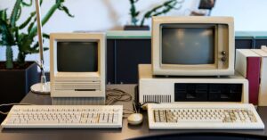 Two vintage desktop computers with monochrome monitors and bulky keyboards displayed on a desk, representing the technological era when the oldest websites were created, with a retro ambiance complemented by a background of potted plants.