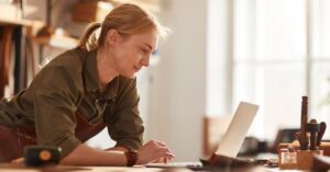 A female solopreneur wearing an apron leans over her workspace, focused on her laptop. She appears to be working in a bright, natural light-filled studio with various tools on the table, reflecting a hands-on, creative work environment.