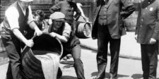 Historic black and white photo of men pouring liquid from a barrel onto the street, observed by onlookers including a police officer.