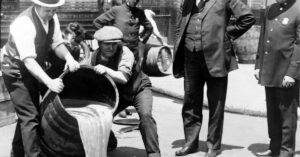 Historic black and white photo of men pouring liquid from a barrel onto the street, observed by onlookers including a police officer.