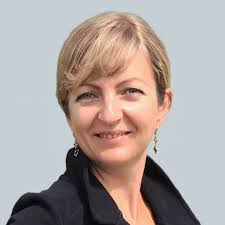 A woman with short blonde hair wearing a black jacket, smiling against a light gray background.