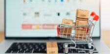 Miniature shopping cart filled with small cardboard boxes placed on a laptop keyboard, representing online shopping and e-commerce. A blurred computer screen in the background displays a shopping website.