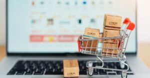 Miniature shopping cart filled with small cardboard boxes placed on a laptop keyboard, representing online shopping and e-commerce. A blurred computer screen in the background displays a shopping website.