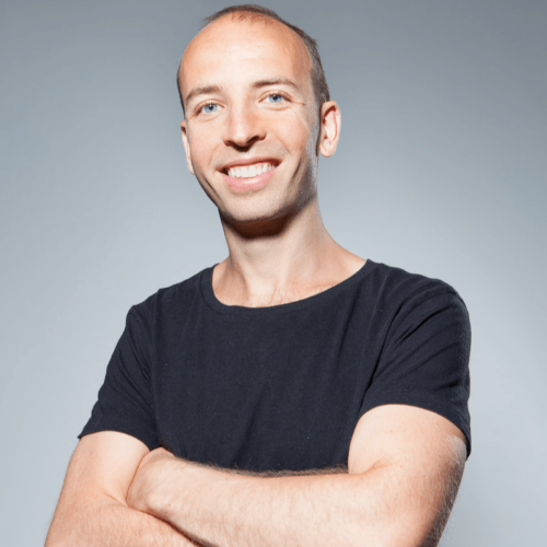 A man with short hair wearing a black T-shirt, smiling with arms crossed against a gray background.