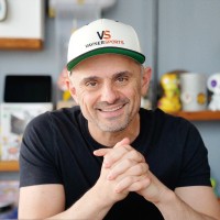 A man wearing a white cap and black T-shirt, smiling with hands clasped, seated in an office setting.