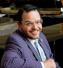 A man wearing glasses and a checkered suit, smiling while seated indoors.