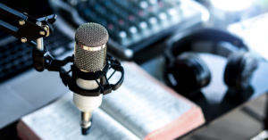 A podcast recording setup featuring a professional microphone on a stand, headphones, an open book, and audio equipment in the background.