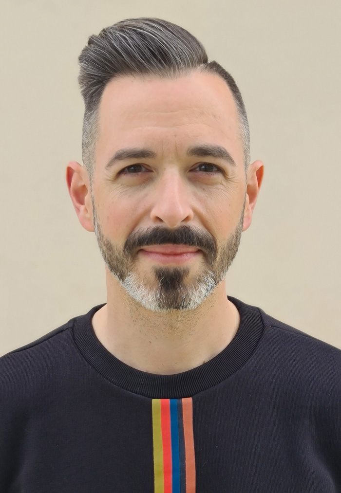 A man with neatly styled hair and a beard, wearing a black shirt with colorful stripes, standing against a beige background.