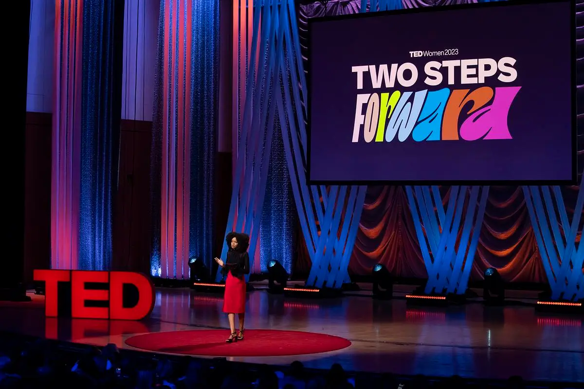 A TED Talk stage with a speaker presenting at TEDWomen 2023. The backdrop features colorful drapes and a large screen displaying the event theme, "Two Steps Forward." The iconic red TED letters are visible on the stage.