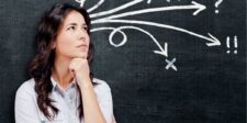 A woman with long dark hair, looking thoughtful, stands in front of a chalkboard with various arrows and symbols pointing in different directions.