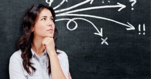 A woman with long dark hair, looking thoughtful, stands in front of a chalkboard with various arrows and symbols pointing in different directions.