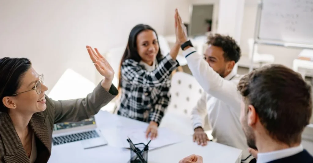 A group of colleagues gathered around a table, smiling and giving high fives in a collaborative office setting.