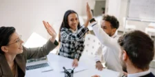 A group of colleagues gathered around a table, smiling and giving high fives in a collaborative office setting.