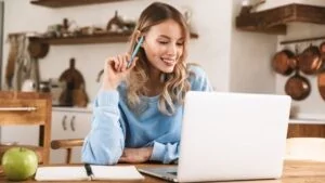 A woman in a cozy kitchen environment smiling while working on her laptop with a pen in hand—capturing focus, comfort, and productive digital work.
