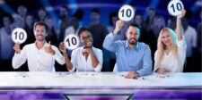 Four judges sitting at a table, each holding up a scorecard with a perfect 10. They are smiling with a blurred audience in the background.