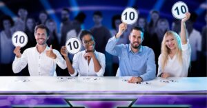 Four judges sitting at a table, each holding up a scorecard with a perfect 10. They are smiling with a blurred audience in the background.