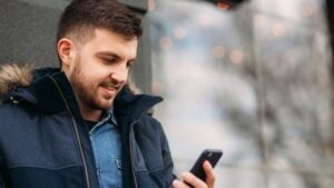 A young man in a jacket checking his phone outdoors, smiling—suggesting access to valuable, timely insights while on the go.