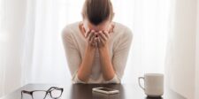 Stressed woman sitting at a table, holding her head in her hands, with glasses, a smartphone, and a white coffee mug on the table in front of her.